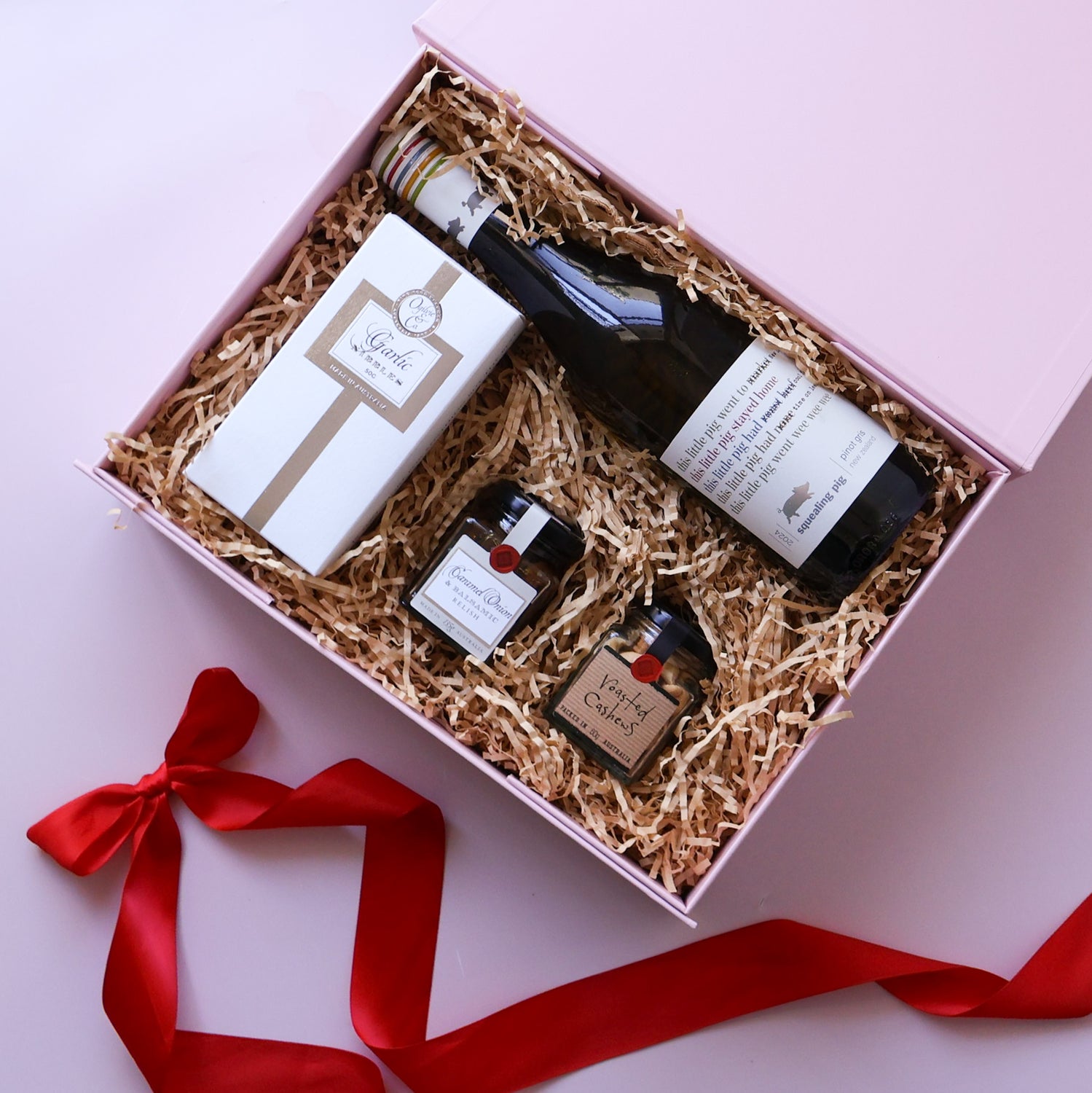 An open flat lay corporate gift hamper tied with a red ribbon, filled with wine, gourmet treats and a wrapped gift on a pink background.