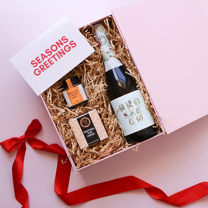 A pink gift box filled with shredded kraft paper contains a dark bottle with a white label, a small orange-labeled item, and a packaged snack. A white card reading “SEASONS GREETINGS” sits in the lid, and a red ribbon is tied around the box on a pink background.