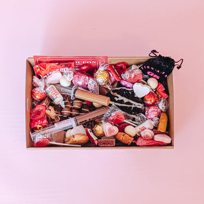 Photograph of valentines gift hamper from above featuring an assortment of treats including two donuts, lindt chocolates, cookies, nutella and biscoff syringes, love coupons and a black velvet pouch containing dirty dice.