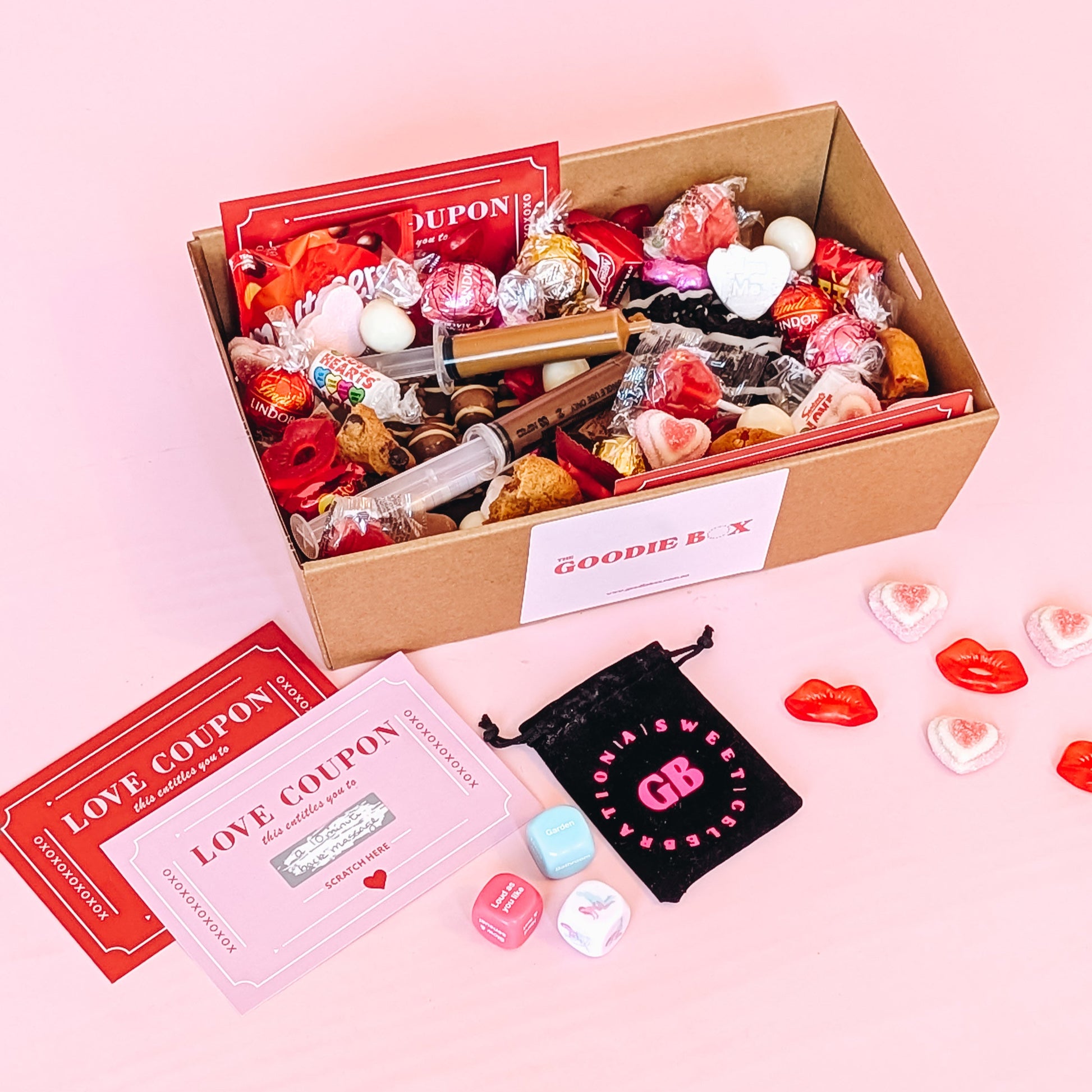 Photograph of valentines gift hamper featuring an assortment of treats including two donuts, lindt chocolates, cookies, nutella and biscoff syringes, love coupons and a black velvet pouch containing dirty dice.