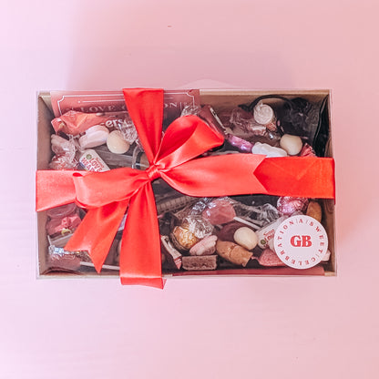 Photograph of valentines gift hamper featuring an assortment of treats including two donuts, lindt chocolates, cookies, nutella and biscoff syringes, love coupons and a black velvet pouch containing dirty dice. Wrapped in a silk red ribbon tied in a bow.