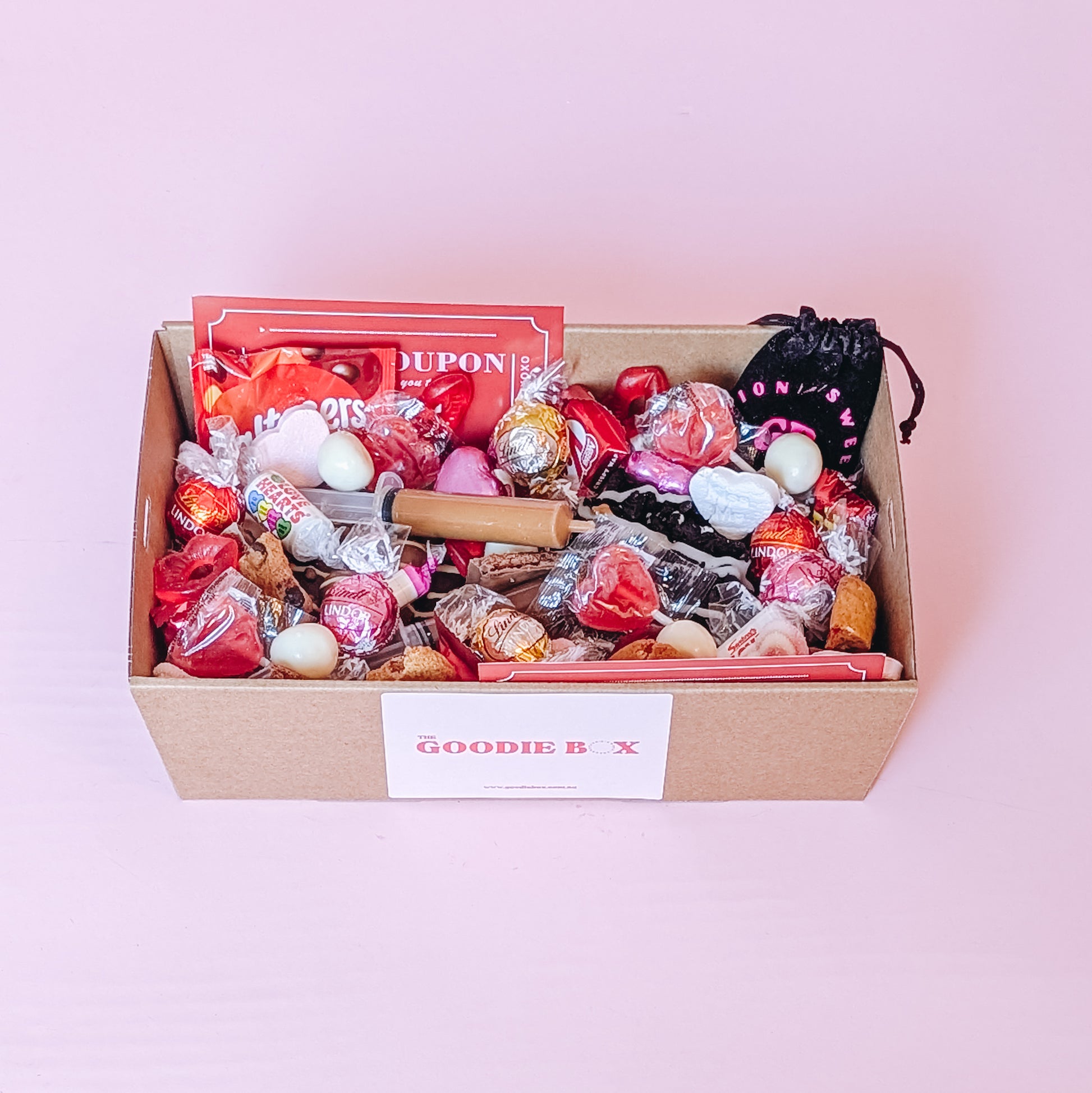 Photograph of valentines gift hamper featuring an assortment of treats including two donuts, lindt chocolates, cookies, nutella and biscoff syringes, love coupons and a black velvet pouch containing dirty dice.