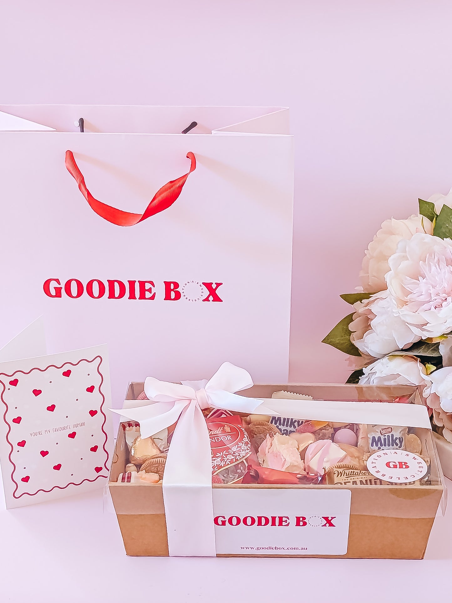Goodie box with a white ribbon, flowers, and a branded bag on a light pink background