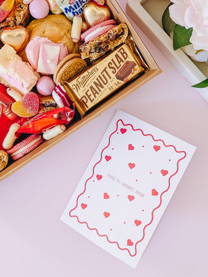 Box of assorted candies with a Whitaker's Peanut Butter Bar, next to a card with heart designs on a pink surface.