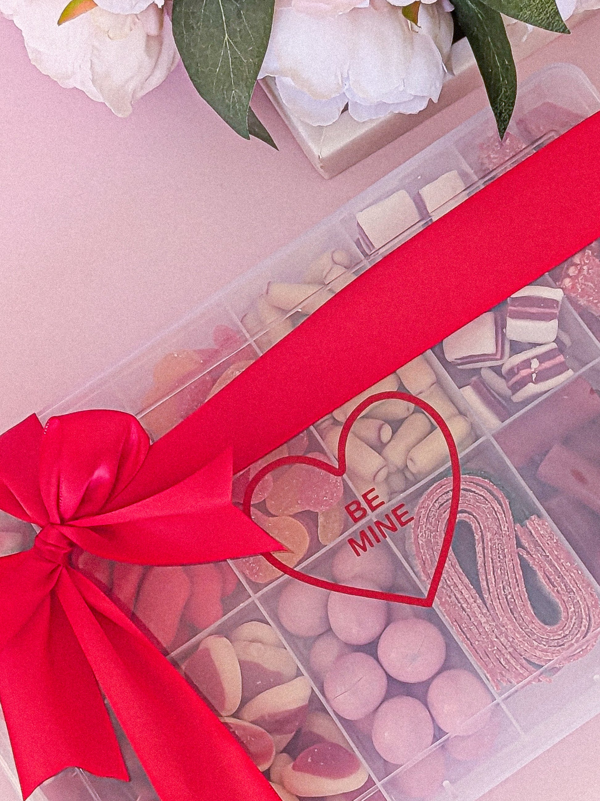 Box of chocolates with a red ribbon and 'Be Mine' heart-shaped tag, surrounded by white flowers.