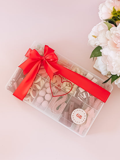 Clear gift box with red ribbon, heart-shaped candy, and GB logo on a light pink background.