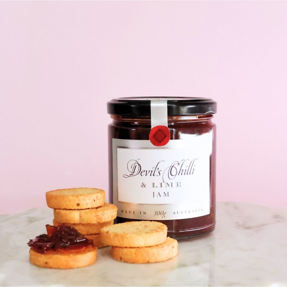 Ogilvie & Co Devil's Chilli & Lime Jam in a 300g jar with a red wax seal, beside stacks of savoury shortbread rounds, one topped with the deep red jam, on marble against a soft pink background.
