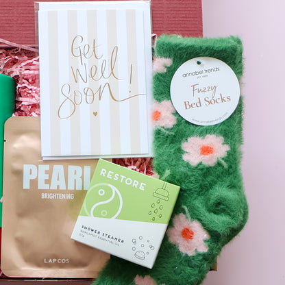 Green fuzzy bed socks with floral patterns, 'Get Well Soon' card, and skincare products on a pink background