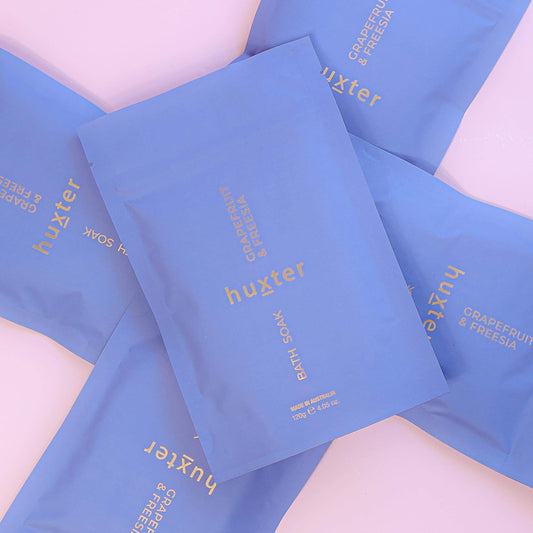 Blue huxter bath soak sachets on a light pink background
