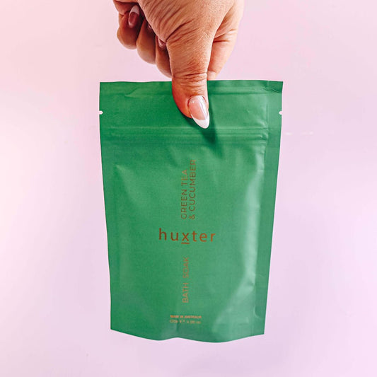 Green Tea & Cucumber Bath Soak package labeled 'huxter' held by a hand against a pink background