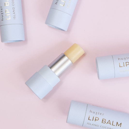 Island coconut lip balm on a pink background, surrounded by blue Huxter lip balm containers.