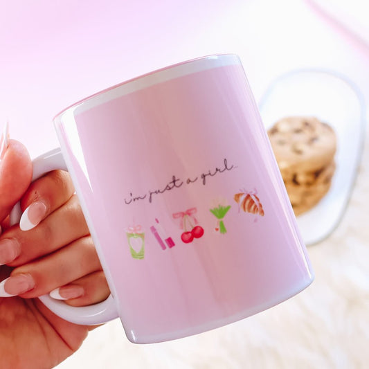 Pink mug reading "im just a girl" with graphics held by a hand, blurred background