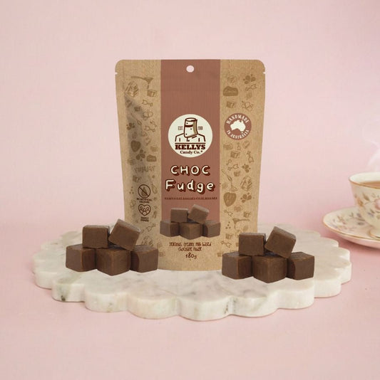 A stand-up pouch of Kelly's Candy Co Choc Fudge is displayed on a scalloped white marble tray with several chocolate fudge cubes arranged in front. A pink background and a partial teacup on the right side complete the scene.