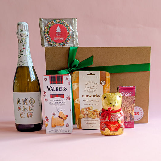 Gift box with bottle of prosecco, buscuits, lindt chocolate and treats on a pink background.