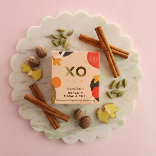 XO Tea Seven Sisters Organic Masala Chai in a peach-toned box, surrounded by cinnamon sticks, cardamom pods, whole nutmeg and fresh ginger on a scalloped marble board.