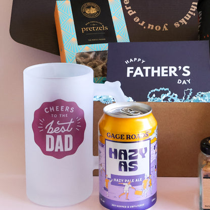 A Father's Day gift set featuring a frosted white mug with a purple 'Cheers to the best Dad' graphic, a can of Gage Road's Hazy As pale ale, and a box of Pretzels, arranged in front of a brown gift box and a 'Happy Father's Day' card.