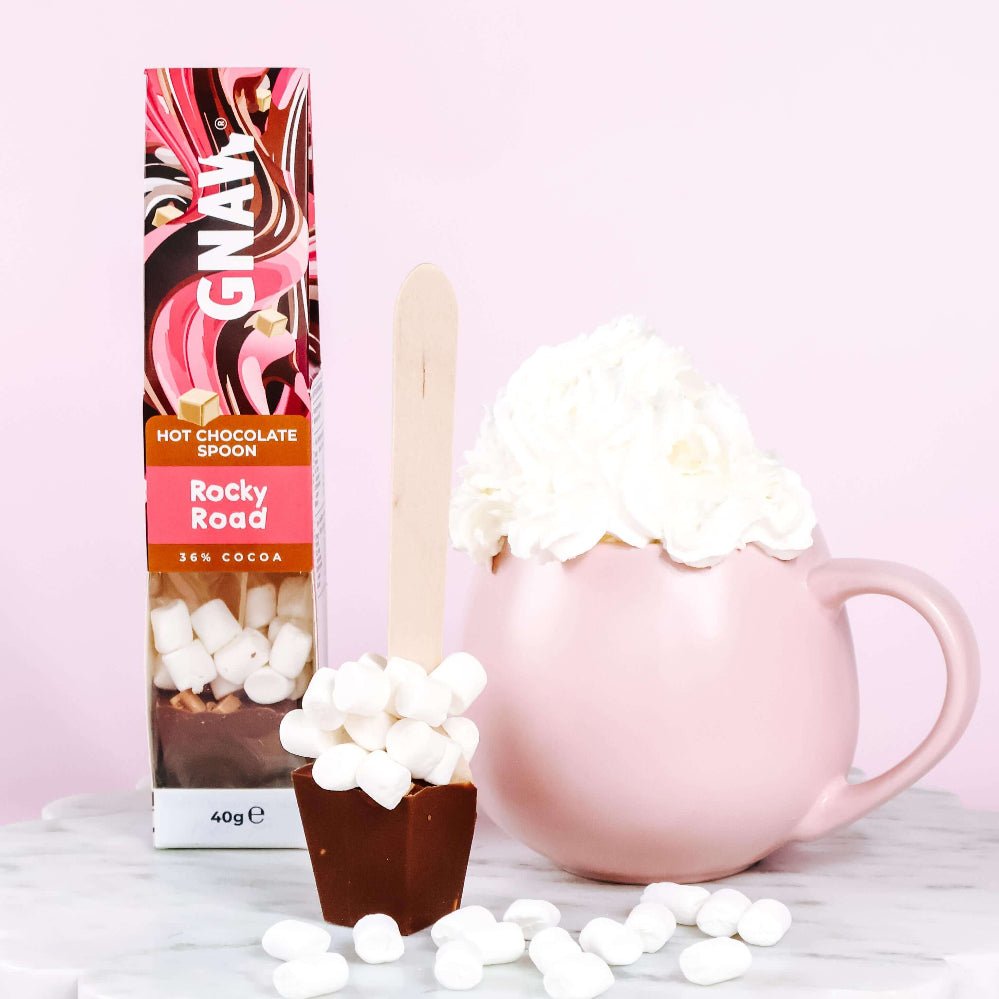 Hot chocolate spoon packaging with rocky road flavor, marshmallows, and a pink mug on a light background.