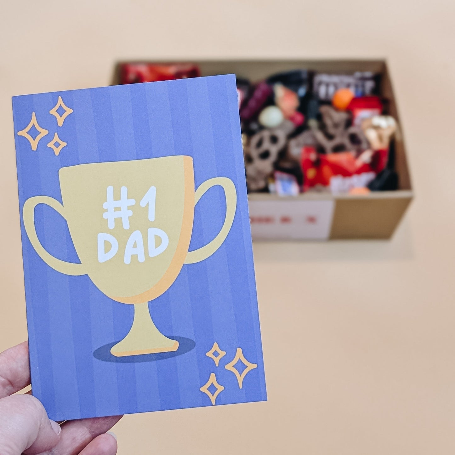 Card with '#1 Dad' trophy design held by a hand, with a gift box in the background.