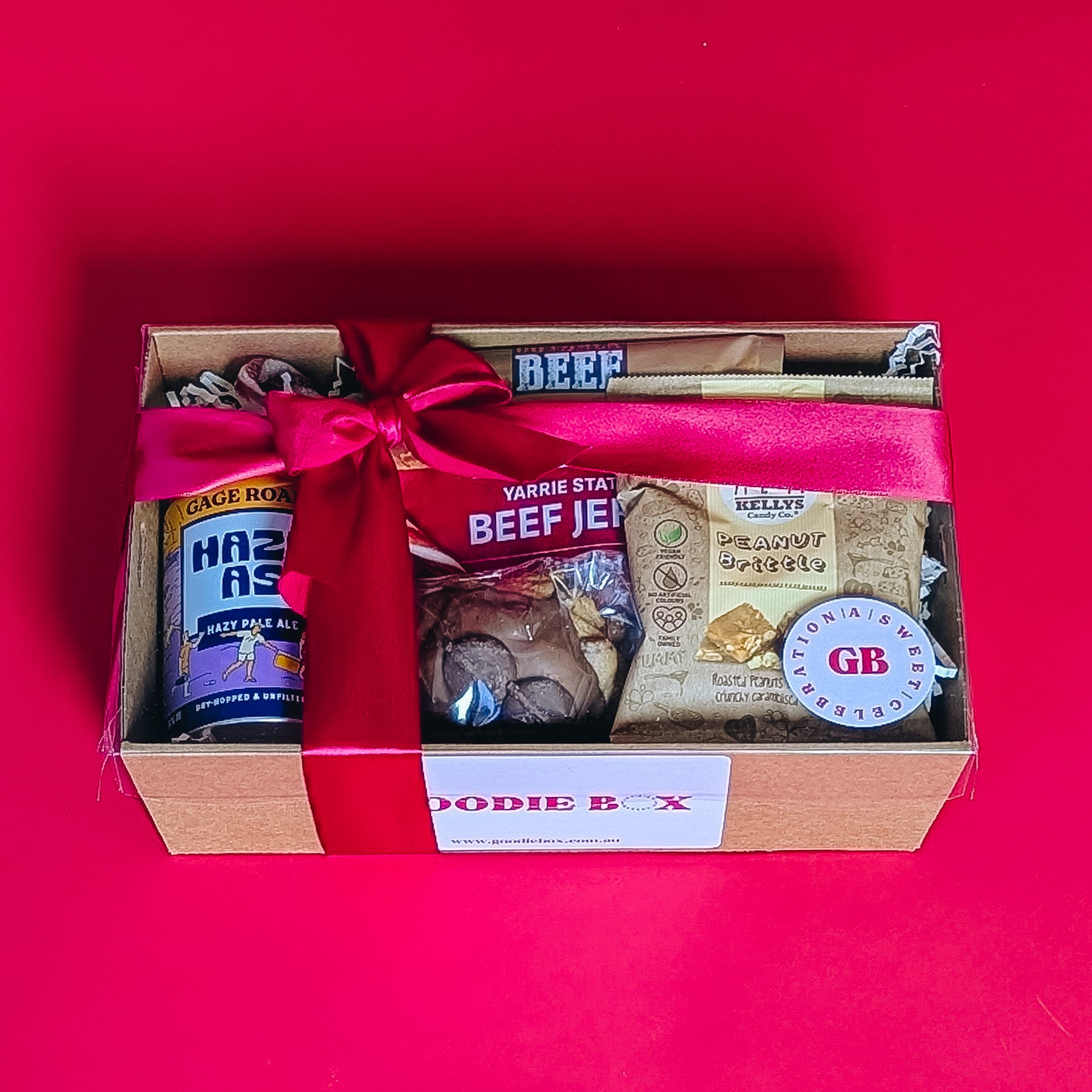 corporate gift box with beef jerkie, pale ale peanut brittle and cookie with a red ribbon on a red background.