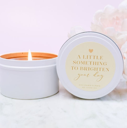 Candle in a white tin with a decorative label reading "a little something to brighten your day" on a light background