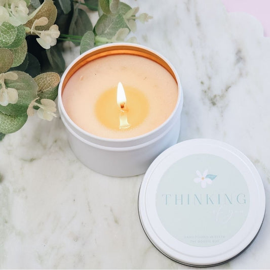 Candle in a white container with a lit flame, reading "thinking of you" surrounded by greenery on a marble surface.