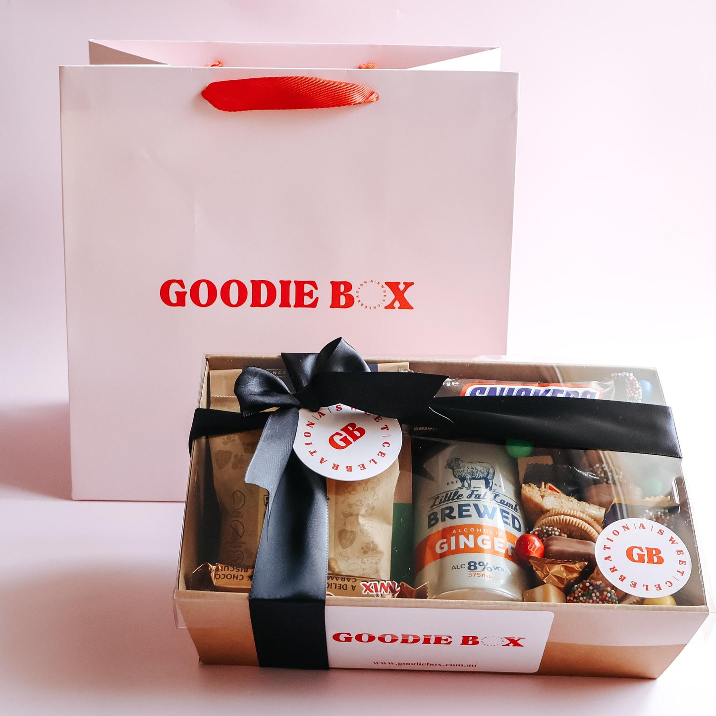 Goodie Box sweet and savoury treat boxfor him with Little Fat Lamb ginger beer, Snickers and treats in branded kraft box with black ribbon and Goodie Box gift bag on pink background Perth