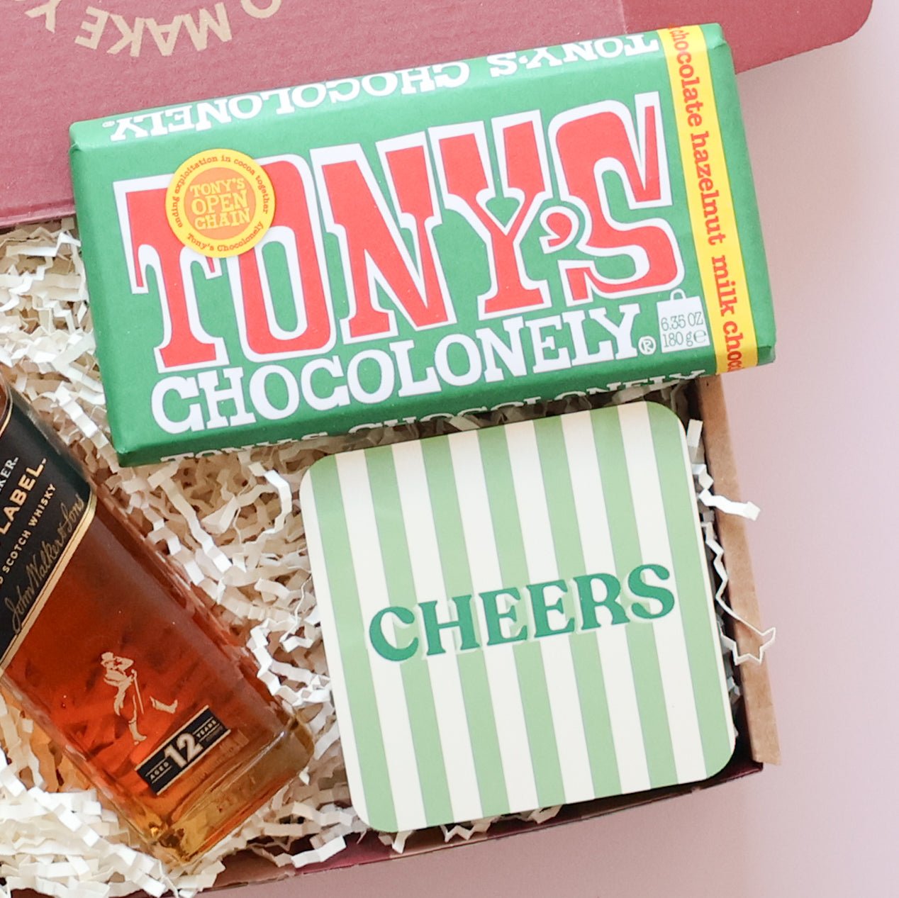 Thoughtful gift for him overhead flatlay showing Johnnie Walker Black Label, Tony's Chocolonely and Cheers stripey coaster in open red gift box