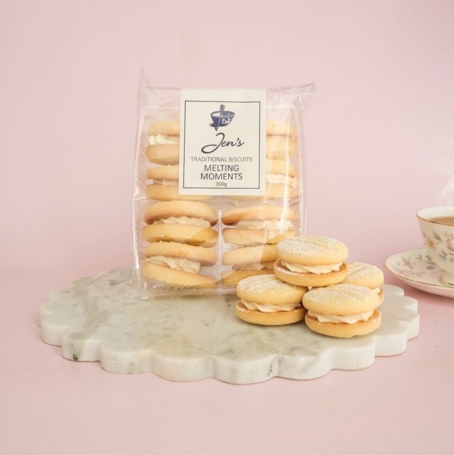 Jen's Traditional Biscuits Melting Moments in a 300g pack, beside a stack of the cream-filled biscuits dusted with icing sugar and a floral china teacup, on marble against a soft pink background.