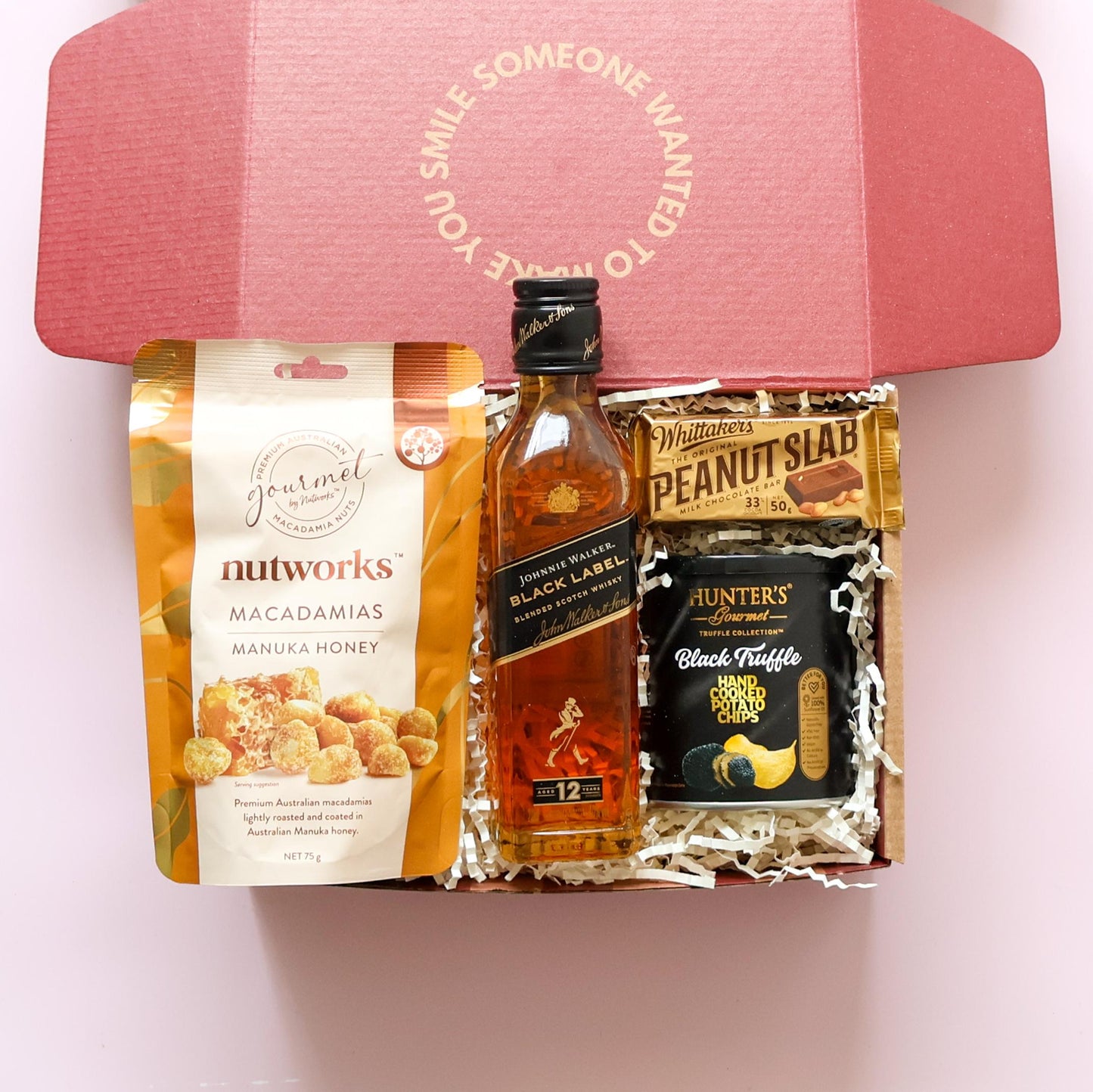 whisky gift box with gourmet snacks including Johnnie Walker Black Label, Whittaker's Peanut Slab, Hunter's Truffle Chips and Nutworks Macadamias on pink background 