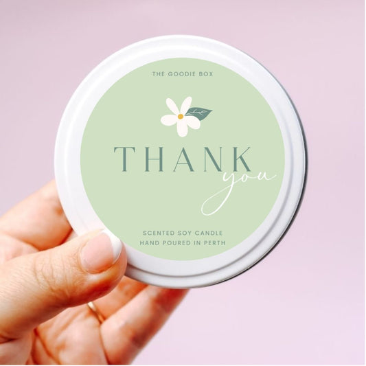 photo of a hand holding a white soy candle tin with green sticker reading "thank you" with white flower